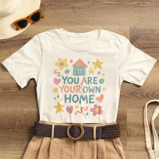 Pastel doodle quote T-shirt featuring "You Are Your Own Home" design by Printify in a naive art aesthetic.