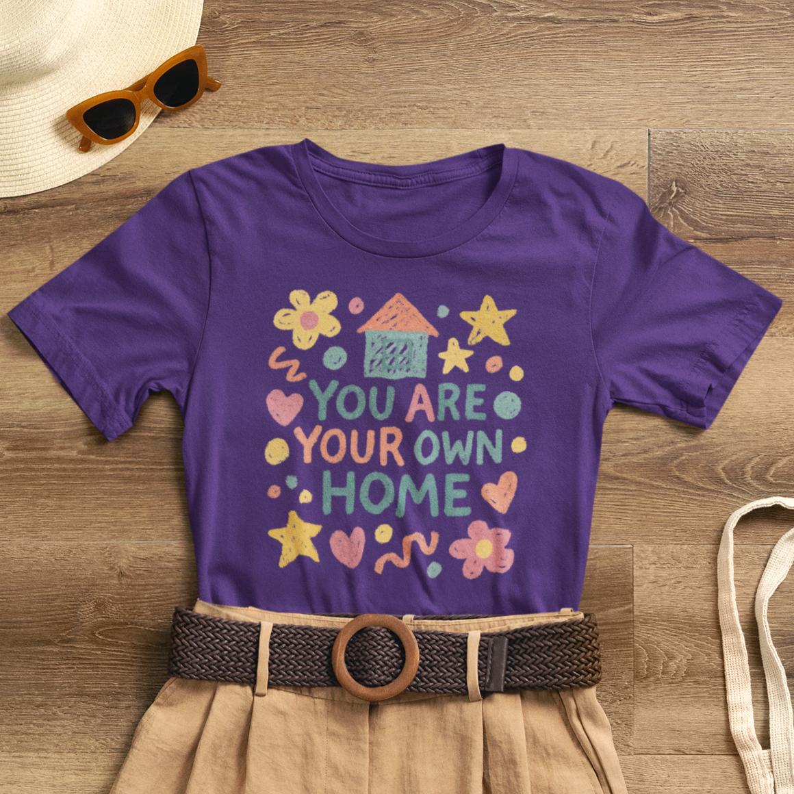 Pastel doodle quote T-shirt featuring "You Are Your Own Home" in naive art style by Printify. Soft, cute, and empowering design.