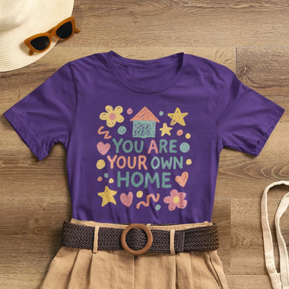 Pastel doodle quote T-shirt featuring "You Are Your Own Home" in naive art style by Printify. Soft, cute, and empowering design.