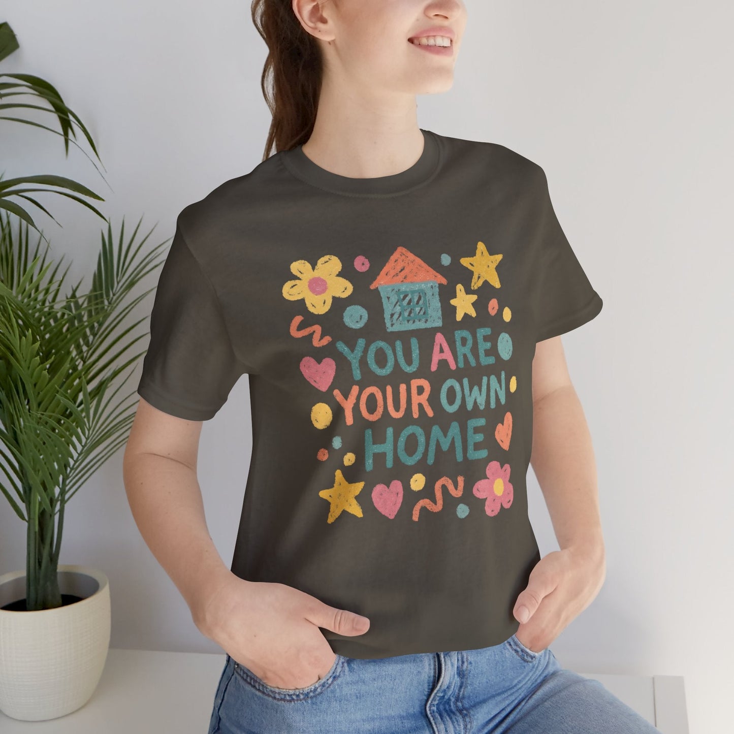 Pastel doodle quote T-shirt featuring "You Are Your Own Home" in naive art aesthetic by Printify. Soft, casual style.